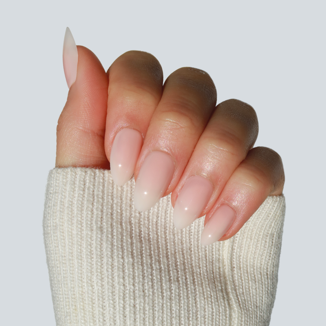 A close-up shot of a hand adorned with Glowie By Her's Daydream Press-On Nails (Short) in a pastel pink shade, polished to a shiny finish. The thumb is slightly up, revealing its design. The hand is partially covered by a beige, ribbed sweater sleeve against a light gray background.