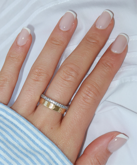 A close-up of a hand adorned with Glowie By Her's Christi nails, showcasing customizable, salon-quality beauty, resting on a white and blue striped cloth. The elegant French tip manicure is enhanced by two rings: a gold band and a silver band studded with small diamonds.