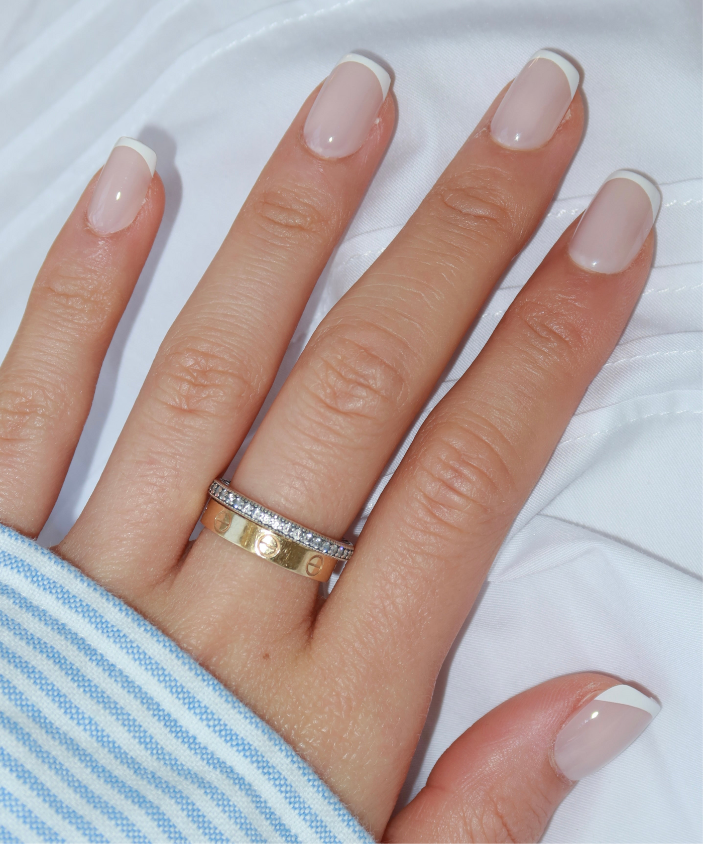 A close-up of a hand adorned with Glowie By Her's Christi Press-On Nails, showcasing salon-quality French tips with white accents on natural pink nails. The hand is accessorized with two rings: a gold band embellished with small diamond accents and another gold band featuring circular engravings, all resting on white and blue-striped fabric.