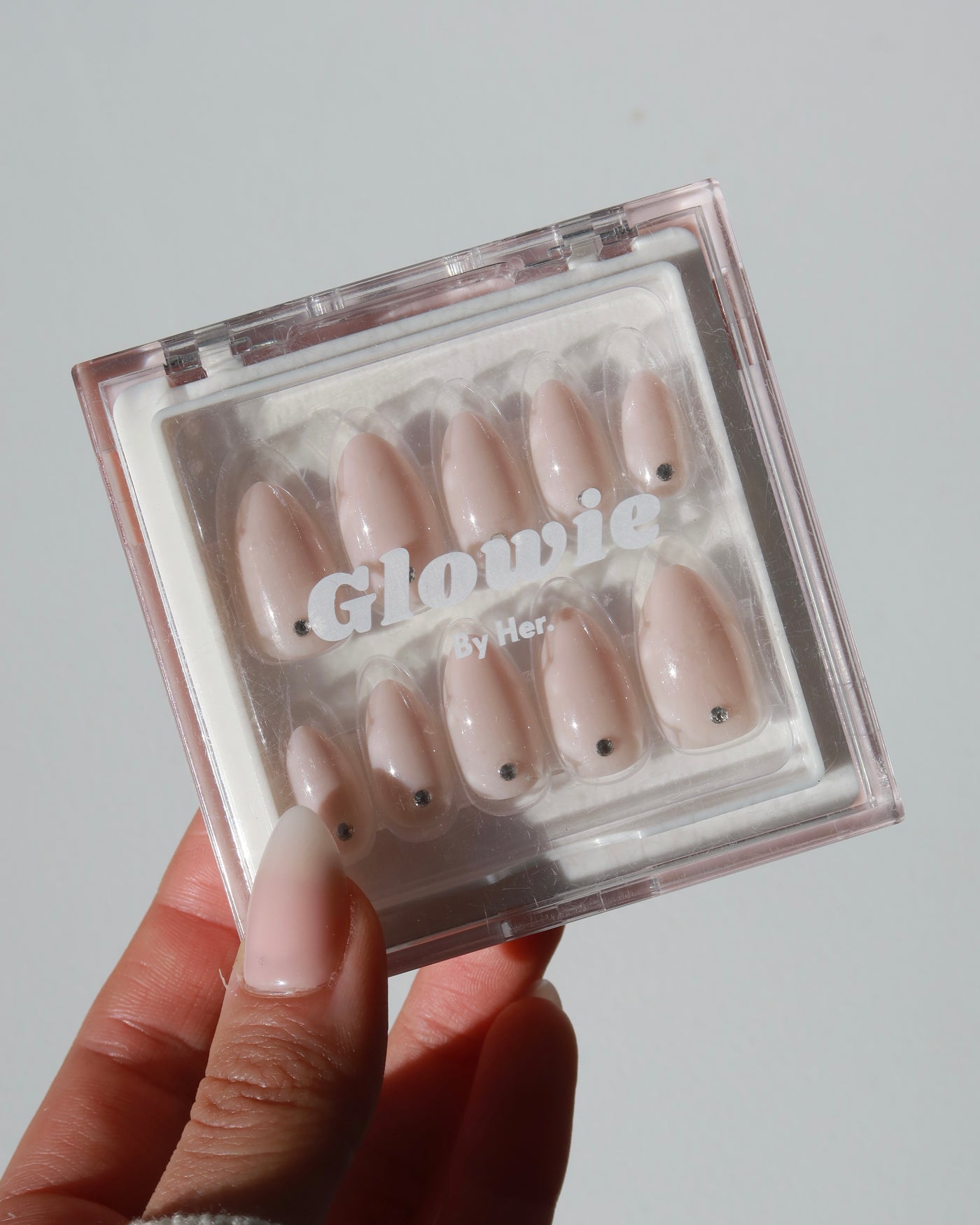 A hand holds a clear plastic case containing ten artificial nails of different sizes. The nails are a pale, nude color with small black dots near the base and have a glossy finish. The text "Glowie By Her" is visible on the case, showcasing the Sweet Talk Collection (now 20% off).