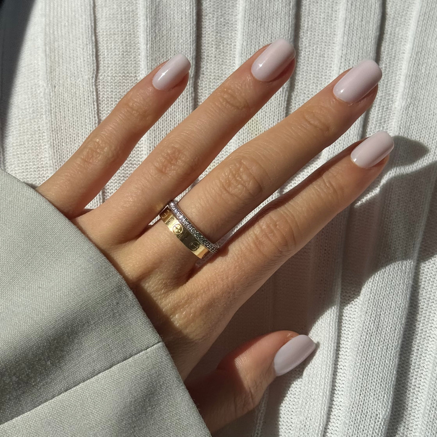 A well-manicured hand with Glowie By Her's Pillowtalk Press-On Nails, featuring salon-quality pink nails, is shown against a white background. The hand is adorned with two rings: one is gold with a small diamond, and the other is silver with intricate detailing. The sleeve of a light gray garment partially covers the wrist.