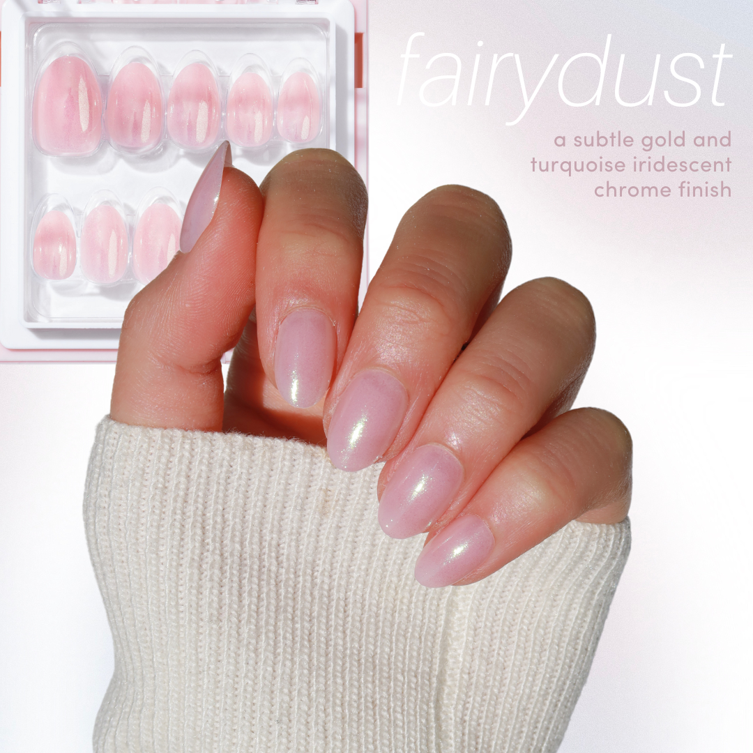 A hand adorned with pink iridescent nails is set against a light background. Next to the hand sits a box from Glowie By Her's 11:11 Collection Vol. 2, complete with a subtle gold and turquoise chrome finish on the "fairydust" nails. The trendy nail designs evoke the mystique of cat eye nails, as the hand is partially wrapped in a cream sweater.