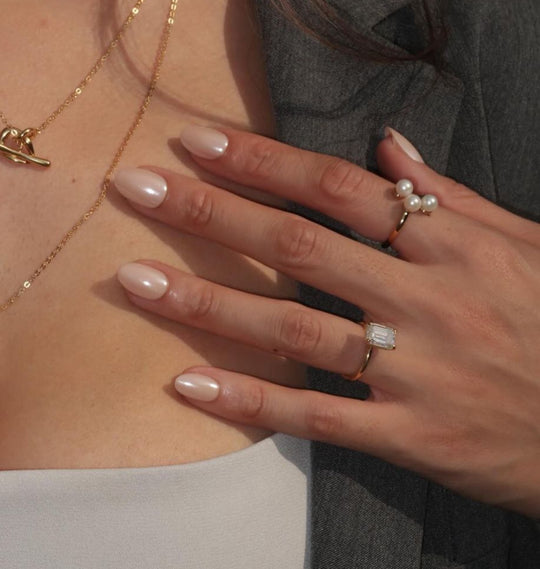 A person with Bubbly, salon-quality nails from Glowie By Her, adorned in a gold necklace, a pearl ring, and a gold ring set with a rectangular gemstone. They are elegantly dressed in a gray blazer over a white top.
