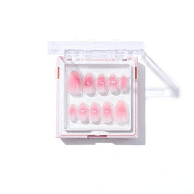 The Made You Blush Press-On Nails by Glowie By Her is a set of salon-quality pink artificial nails with a blush nail base and 3D gel heart accents, neatly arranged in a white, rectangular plastic case with a clear lid. The nails come in various sizes to fit different fingers, and the open case showcases the beautifully designed nails inside.