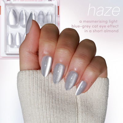 A hand showcasing manicured nails in a shimmering light blue-grey, styled in a short almond shape, is set against a white background. Accentuated by a cozy beige sweater, the nails feature a subtle cat eye effect. In the background is the "11:11 - Full Collection (35% Off)" nail set by Glowie By Her, labeled "haze.