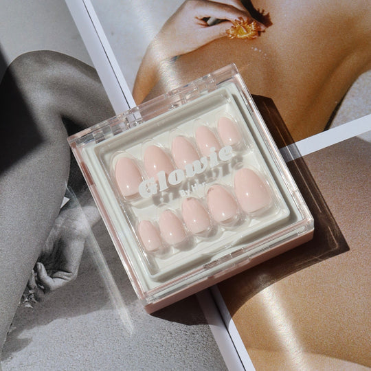 A box of salon-quality pastel pink press-on nails labeled "Sofia" from Glowie By Her. rests on an open magazine showcasing black and white and color images of a woman's body. The sunlight casts soft shadows, adding a warm tone to the scene.