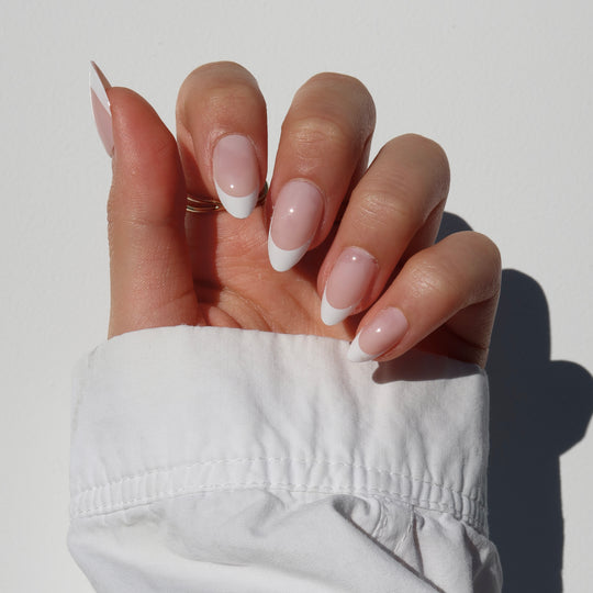 A hand featuring the 'That girl' nails by Glowie Co., with salon-quality French nails that boast long, natural pink bases and crisp white tips, is set against a white background. The wearer has on a white, long-sleeved shirt that complements the 100% customizable elegance of this timeless manicure.