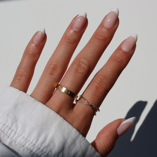 A well-groomed hand featuring Glowie Co.'s ‘That girl’ salon-quality French nails, showcasing elegant white tips. The hand is adorned with two rings: a gold band with engraved details and a thin, delicate ring. The sleeve of a crisp white shirt partially covers the wrist.