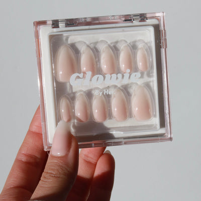 A hand holds a clear plastic box containing a set of Daydream Press-On Nails (Short) in varying sizes. The nails are a natural pink shade. The text on the box reads, "Glowie By Her.