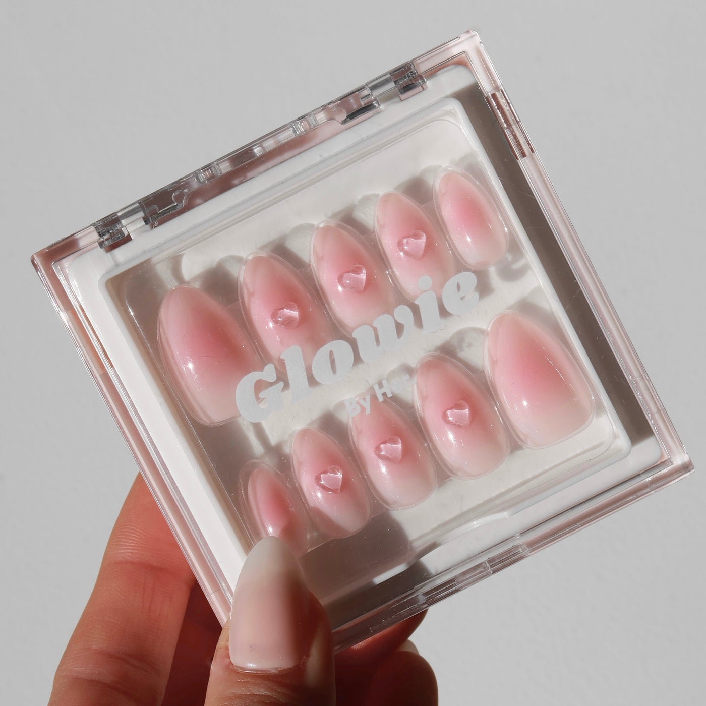 A hand is holding a clear plastic box containing a set of salon-quality pink press-on nails with small 3D gel heart accents. The brand name "Glowie By Her" and the product name "Made You Blush Press-On Nails" are visible on the box lid.