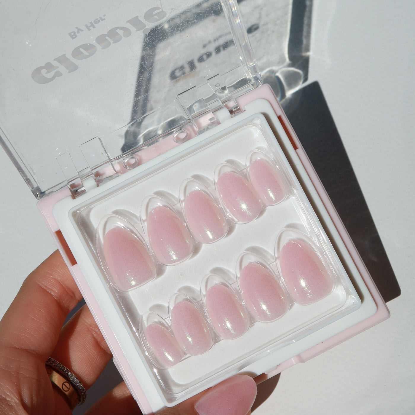 A hand holds a clear plastic case containing a set of Fairydust Press-On Nails by Glowie By Her, featuring salon-quality, pink almond-shaped artificial nails. The sunlight casts a shadow of the case and nails on the white surface in the background, showcasing their iridescent chrome finish.