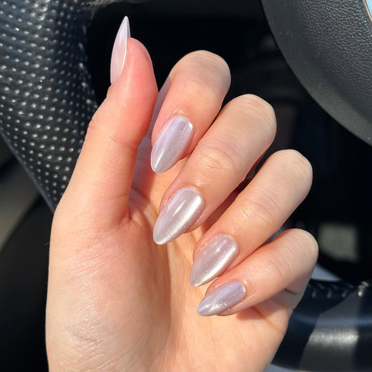 A hand with long, almond-shaped nails, adorned in Glowie By Her's ASAP shimmering pink cat-eye design, rests on a black textured car steering wheel.