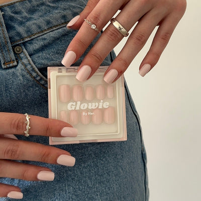 A person with light pink manicured nails holds a set of salon-quality false nails labeled "Pillowtalk" by Glowie By Her. They are wearing jeans and have silver rings on their fingers, showcasing customizable nails perfect for any occasion.