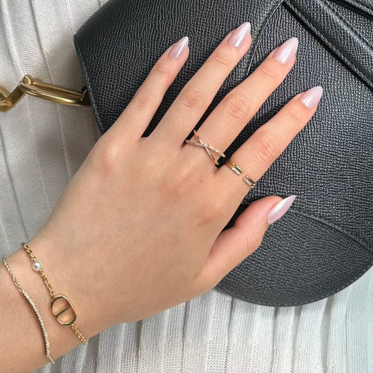 A hand featuring Hailey Re-Edition press-on nails by Glowie By Her, showcasing pink chrome glitters, is adorned with gold rings and bracelets, resting on a textured black purse.