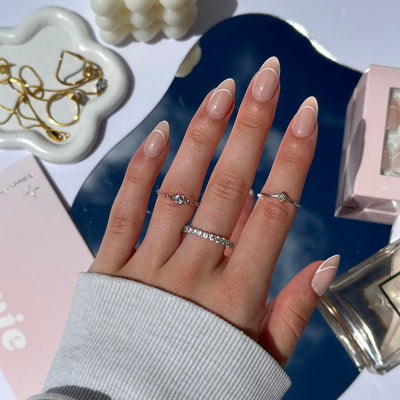 A hand featuring salon-quality nails and adorned with three silver rings rests on a reflective blue surface. Nearby, gold jewelry, a candle, and perfume bottles from Glowie Co.'s Halo (Re-edition) are partially visible. The white and pink background enhances the scene's elegant and refined atmosphere.