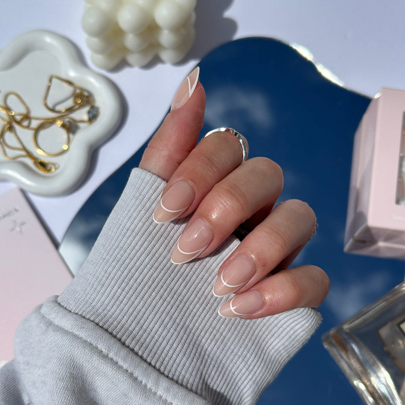 A hand adorned with the elegant nude and white French manicure of Glowie Co.'s Halo (Re-edition) salon-quality nails rests on a reflective surface, partially covered by a gray sleeve. Nearby, a decorative dish with gold jewelry and a bubble-shaped white candle adds to the ambiance.