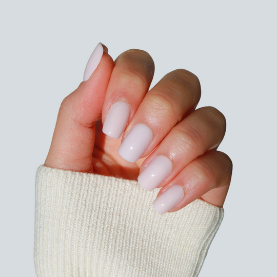 A hand with neatly manicured nails painted in the soft pastel pink shade called Pillowtalk by Glowie By Her holds a beige knitted sleeve. The salon-quality nails are glossy, and the cozy sleeve enhances the image, set against a plain light gray background.