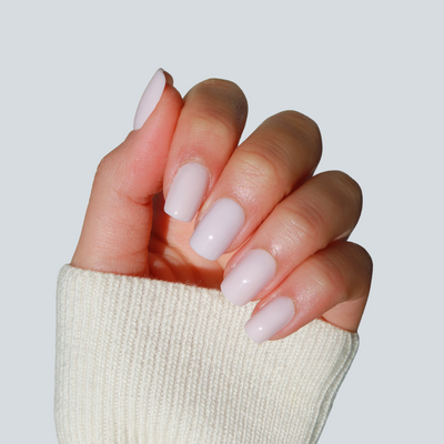 A hand with neatly manicured nails adorned in a soft, pastel pink color using Pillowtalk Press-On Nails by Glowie By Her is shown against a light blue background. The person is wearing a white, ribbed sweater with the sleeve partially covering the wrist. Achieve salon-quality nails at home effortlessly with this easy-to-use nail kit from Glowie By Her.