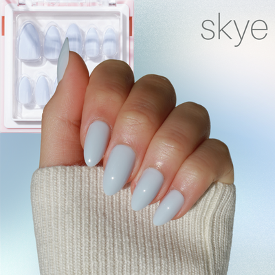 A hand with long, salon-quality, light blue almond-shaped nails glides against a soft gradient background. The sleeve of a cream-colored sweater gently covers the wrist. In the top left, a set from Glowie By Her's Blue Mood collection is packaged neatly, featuring "skye" in stylish text on the top right and highlighting its new release at 25% off.