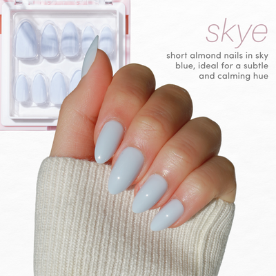 A hand with short almond-shaped nails painted in a sky blue color holds a cream-colored knit fabric. In the background, a set of sky blue nail tips from the Essentials Collection 2.0 by Glowie By Her are displayed in a plastic case. The image text reads: "skye - short almond nails in sky blue, ideal for a subtle and calming hue." Enjoy 20% off now!