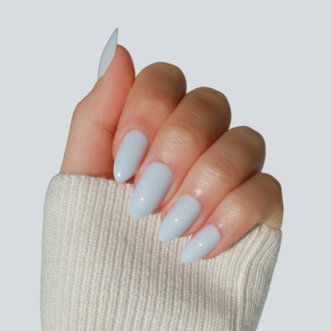 A hand with long, almond-shaped, salon-quality nails painted in the light greyish-white shade of Skye Press-On Nails by Glowie By Her is shown against a plain background. The hand is partially covered by a light-colored, ribbed sweater sleeve pulled over the wrist.