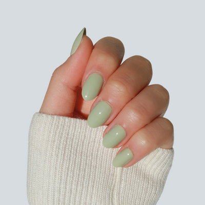 A hand with a pale green, glossy manicure featuring So Sage Press-On Nails by Glowie By Her is held up against a light gray background. The fingers are slightly curved, and the person is wearing a cream-colored, ribbed sleeve that partially covers the hand. These salon-quality nails are 100% customizable and always vegan & cruelty-free.