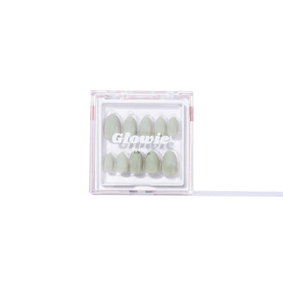 A clear rectangular box from Glowie By Her, containing the So Sage Press-On Nails—a set of salon-quality, pale muted green false nails. The box has partially visible markings or text and these 100% customizable nails are vegan and cruelty-free. The background is white.