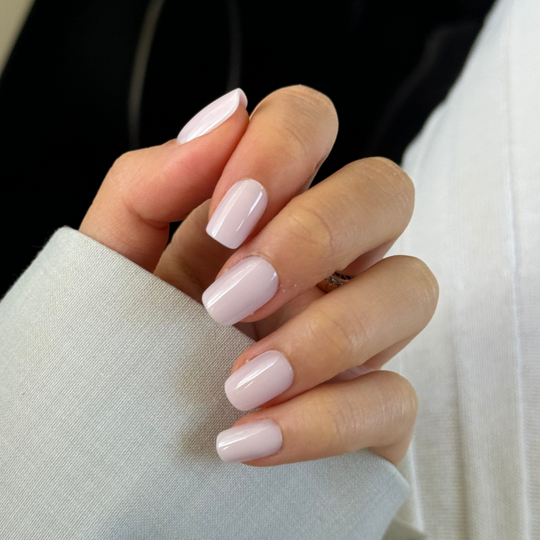 A hand with neatly manicured, salon-quality nails painted in Pillowtalk, a pale pink shade from Glowie By Her. The fingers are slightly curled, and the person is wearing a light-colored long-sleeve garment. The dark background accentuates the elegance of these beautifully crafted nails.