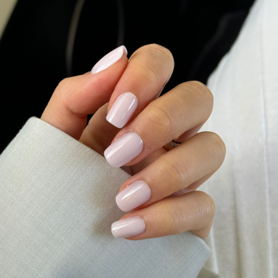 A close-up of a hand with neatly manicured, salon-quality Pillowtalk Press-On Nails by Glowie By Her, painted in a light pink shade. The hand is resting on a light-colored fabric, possibly a sleeve, and the background is softly blurred, enhancing the focus on the nails.