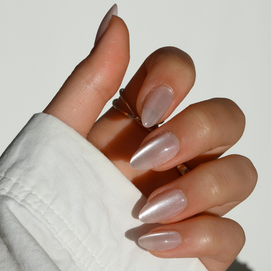 A hand embellished with Glowie By Her's ASAP almond-shaped pink cat-eye nails displays a glossy, translucent finish. Set against a light background, the image emphasizes salon-quality elegance, with a white sleeve appearing at the bottom of the scene.