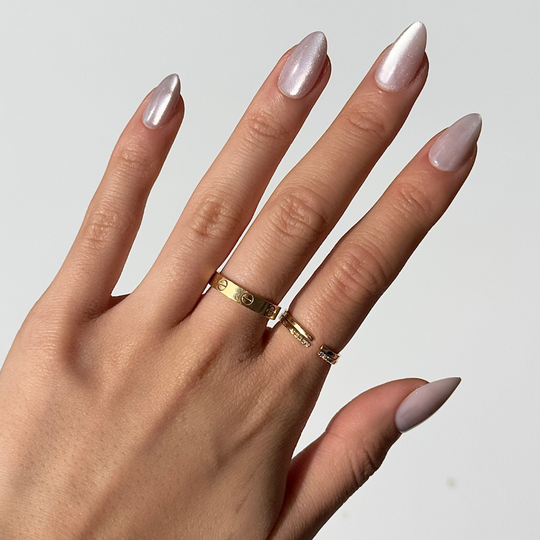 A hand adorned with long, glossy, pink cat-eye nails from Glowie By Her's ASAP collection is featured. The person wears three gold rings on their fingers against a white background.