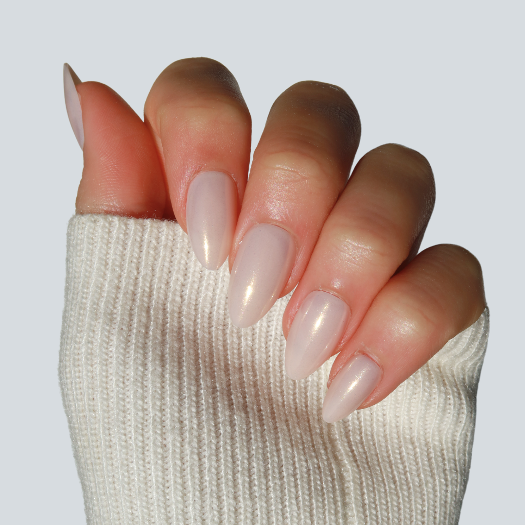 A close-up of a hand adorned with Glowie By Her's Vanilla Press-On Nails in a glossy baby pink shade. The salon-quality nails are partially covered by the sleeve of a cozy, white ribbed sweater, creating a soft and elegant look against a light grey background.