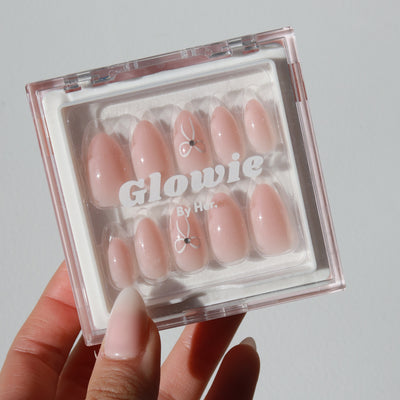 A hand holds a clear plastic case containing a set of glossy, baby pink "Cutie Press-On Nails" in an elegant almond shape. The case has "Glowie By Her" written on it in white letters. Two tiny cherry blossom designs are visible on some of the customizable nails.