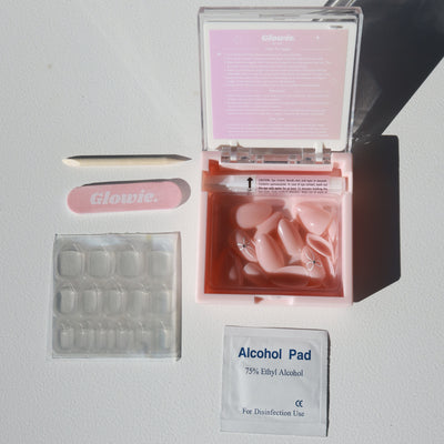 The image showcases a set of nail care items, including a pink box labeled 'Glowie By Her' containing the 'Cutie Press-On Nails' in a baby pink almond shape, accompanied by a pink nail file, wooden cuticle stick, adhesive tabs, and an alcohol pad for disinfection. Everything is arranged neatly on a white surface.